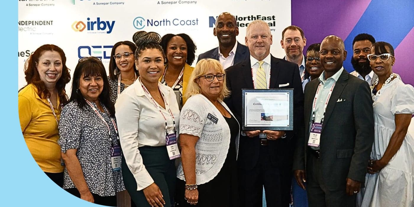 A group of people standing in front of a white backdrop. A person in the middle holds a framed award; they wear a black suit and tie.